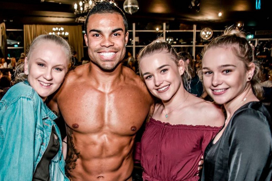 A smiling topless man posing with three young women in a chandelier-lit venue