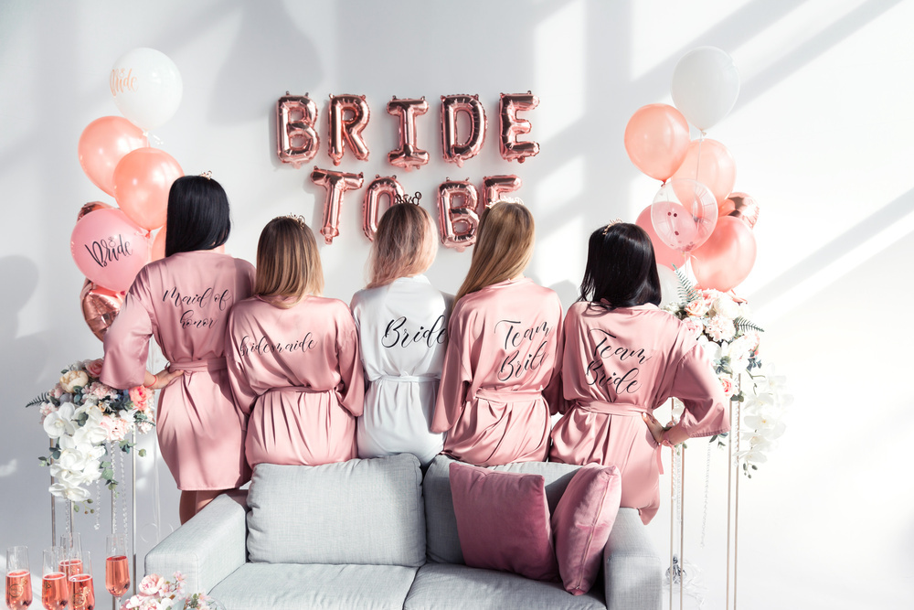 Five friends in matching pink robes seen from behind, beside Bride To Be balloons
