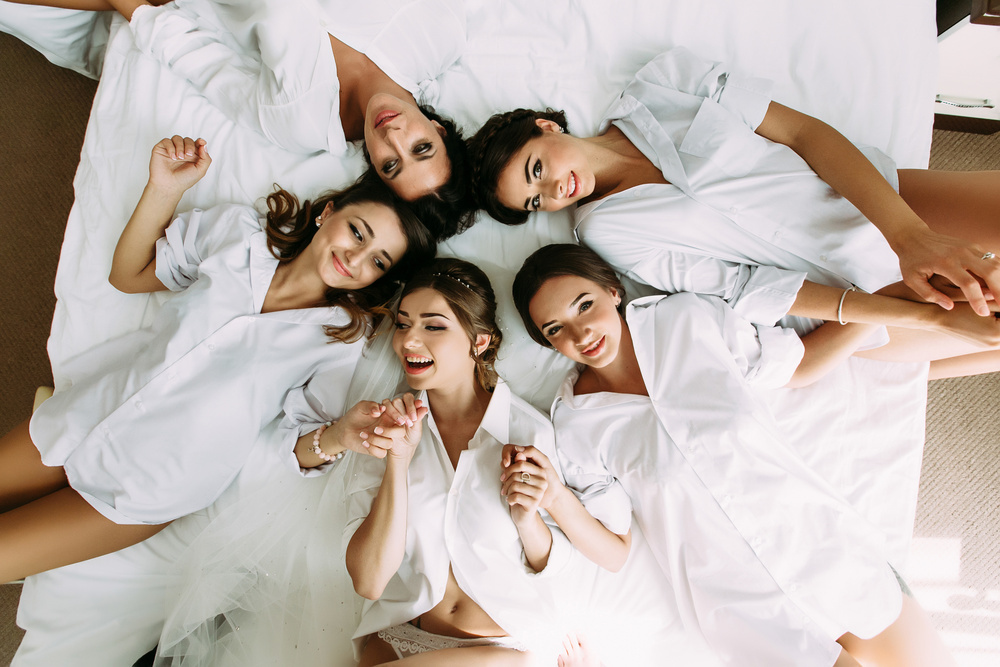 A circle of friends in white robes lying head to head on a bed, laughing up at the camera