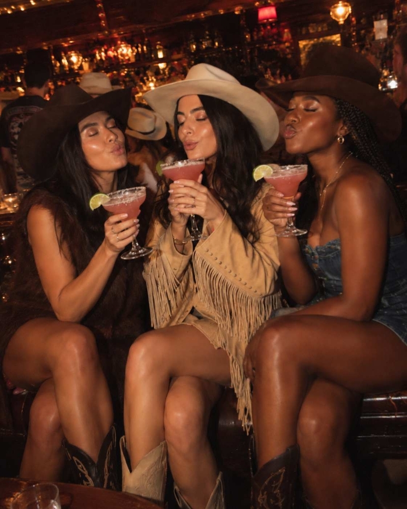 Three hens in cowboy hats toasting margaritas at night