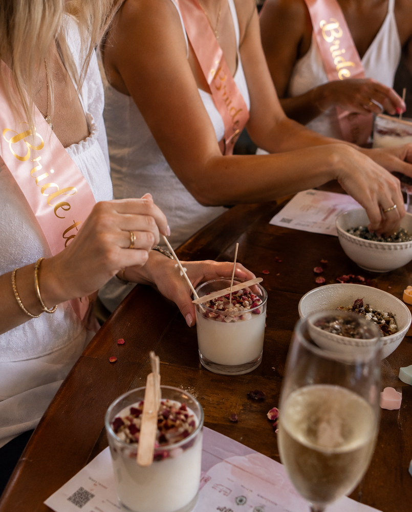 Hens in sashes decorating freshly poured candles with dried petals beside a glass of bubbles