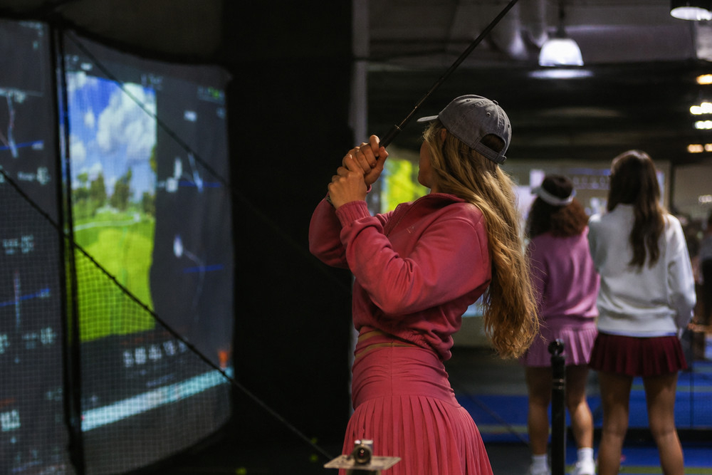 A hen in a pink pleated skirt takes a swing at a golf simulator screen