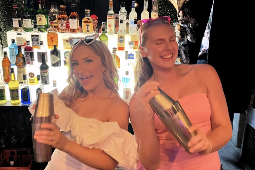 Two hens laughing as they shake cocktail shakers in front of a lit back bar