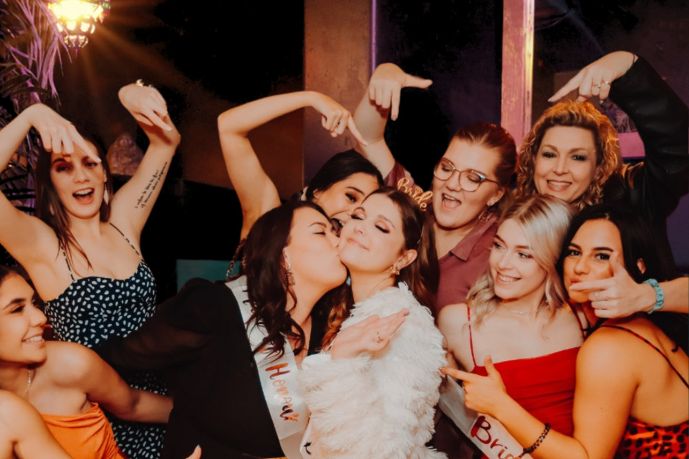 A bride in white feathers getting a kiss on the cheek as her bridesmaids in sashes pose around her