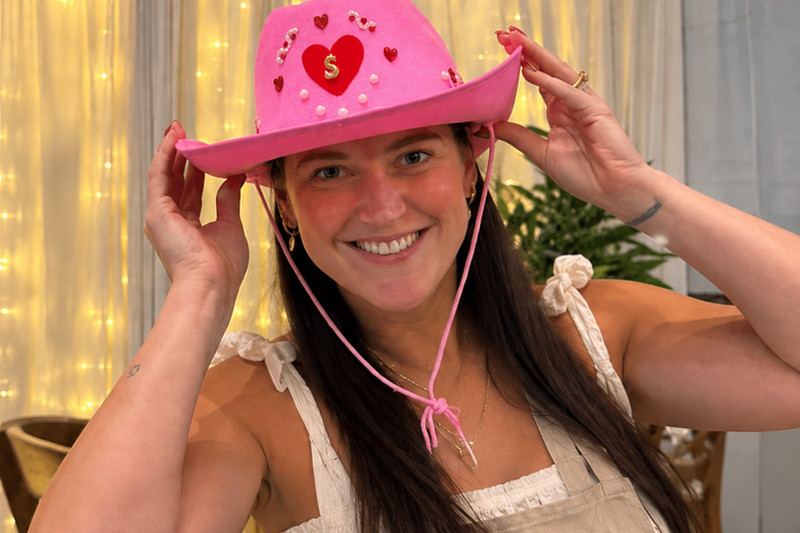 A smiling woman in an apron showing off her decorated pink cowboy hat