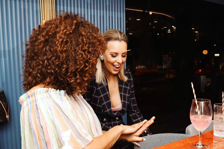 Two friends at a nail bar, one showing off her fresh nails, a pink drink beside them