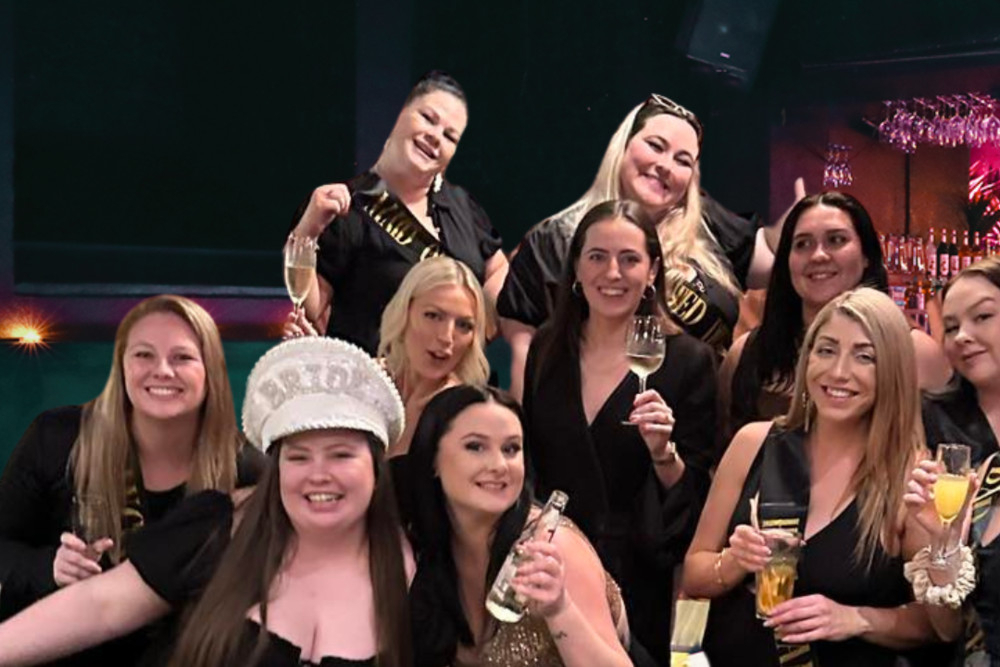 A hens group in black with the bride in a white hat raising drinks in a lounge