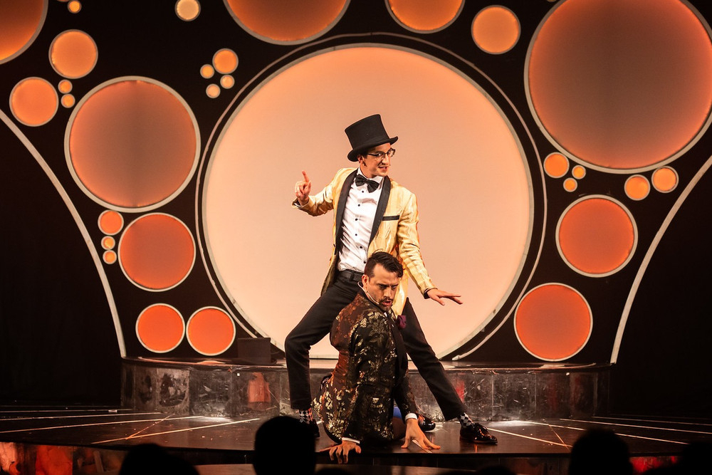 Two cabaret performers in top hats striking a pose on a stage framed by glowing circles
