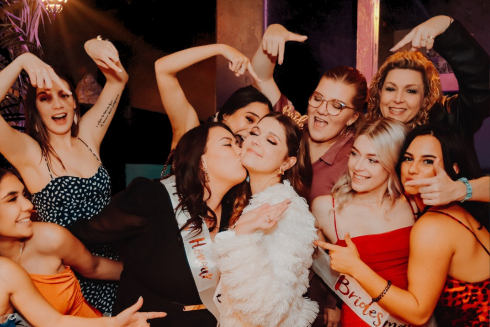 Friends in going-out dresses dancing and pulling poses around the bride, one kissing her cheek