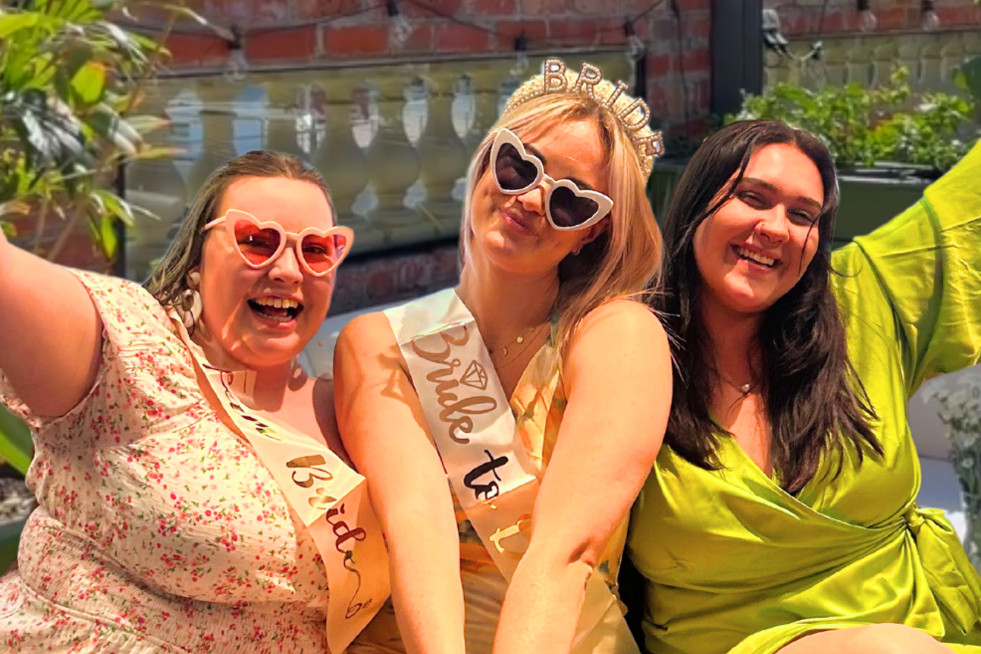 The bride to be in a sash and heart sunglasses laughing with two friends on a sunny city rooftop