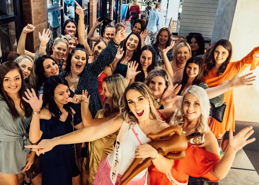 The bride in a sash cheering with her big crew of friends, arms up, on a night out