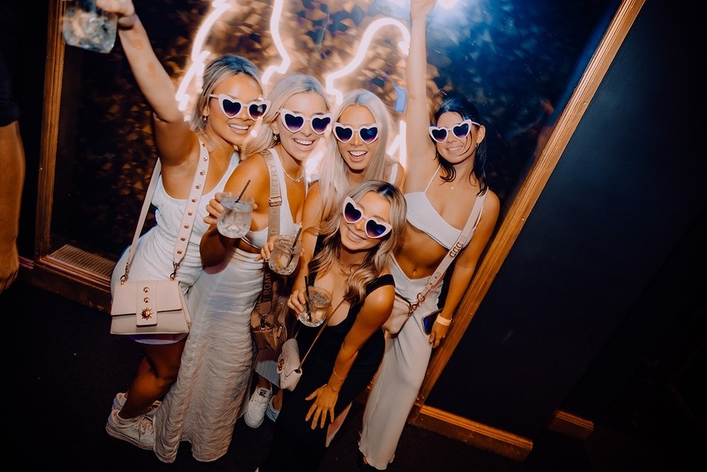 Friends in white outfits and heart sunglasses raise their drinks on a night out