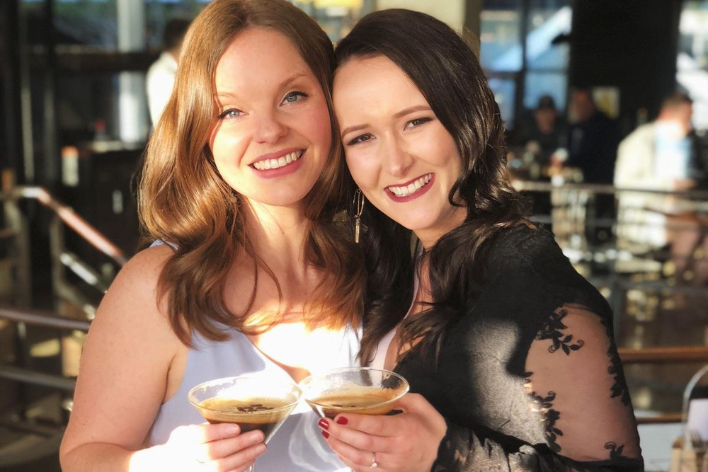 Two friends cheek to cheek toasting with cocktails, one wearing a bride sash