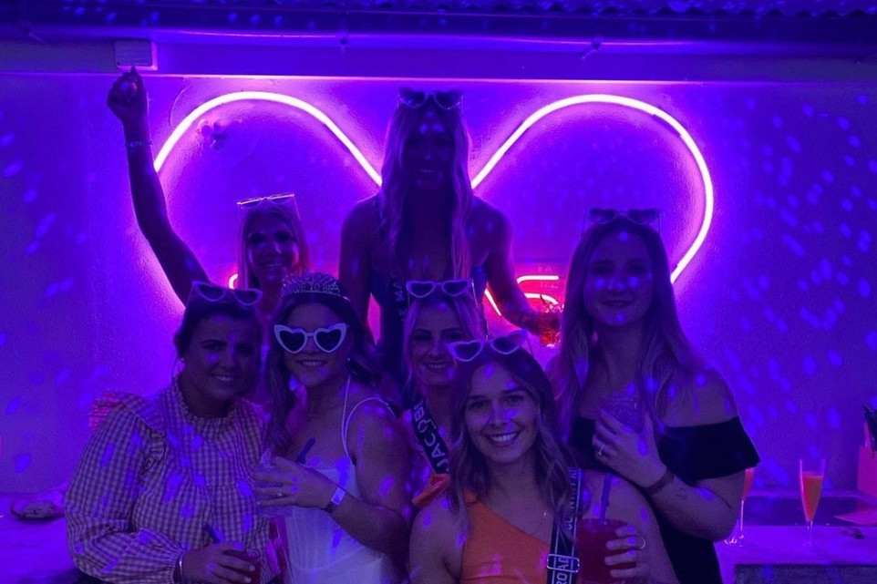 A hens group in heart sunglasses and sashes under a glowing neon heart