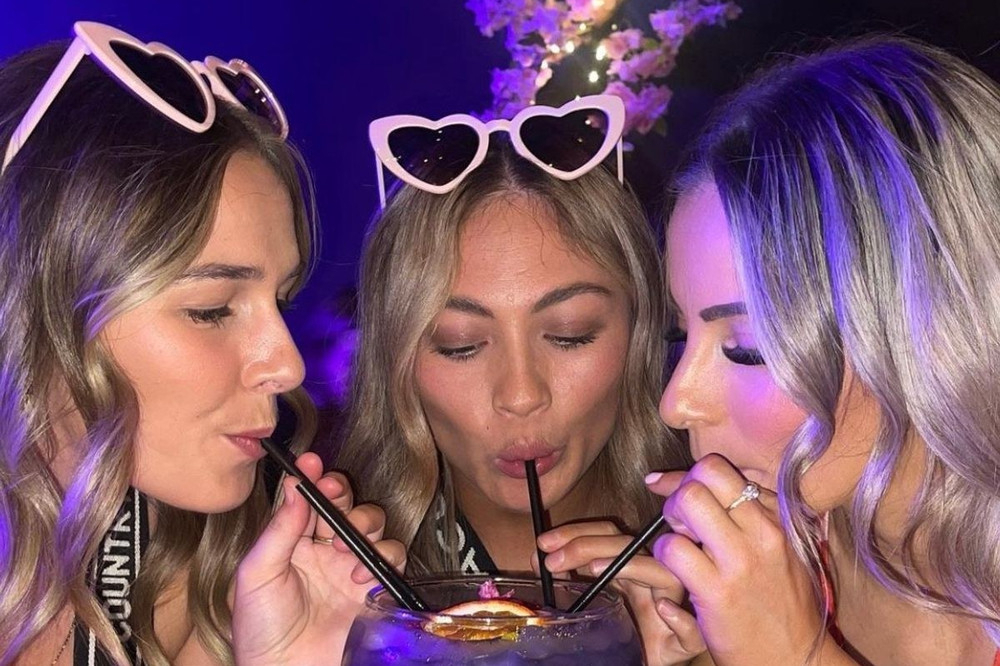 Three friends in heart sunglasses sipping one cocktail through straws under purple light