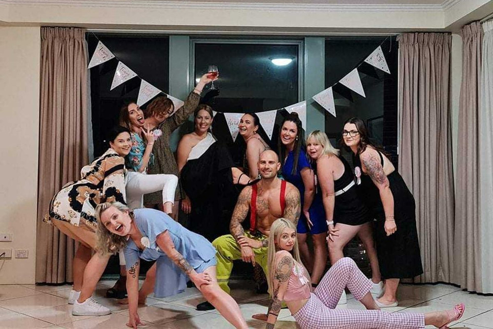 A hens group striking playful poses with their shirtless waiter under white bunting on an R&B hens night