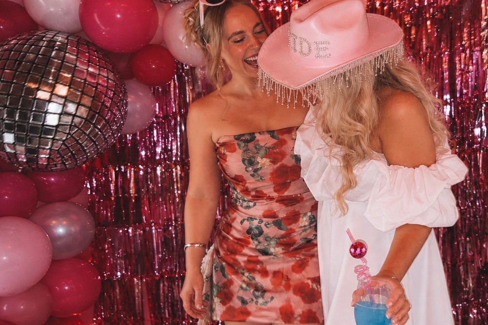 The bride in a pink cowgirl hat laughing with a friend beside pink tinsel and disco balls