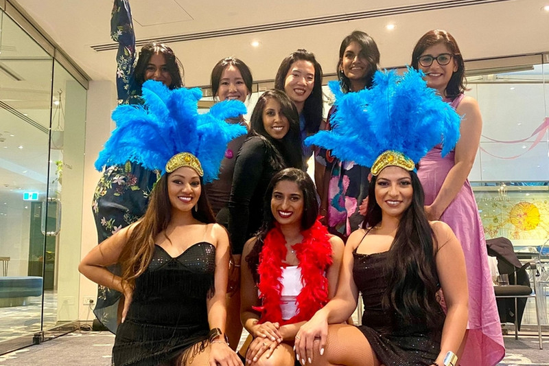 A hens crew in black dresses posing with big blue feather headdresses and a red boa
