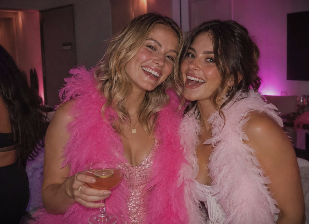 Two friends in pink and white feather boas laughing with cocktails in a pink-lit room