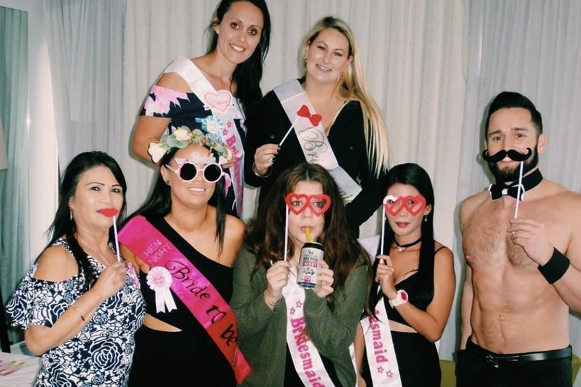 Hens in sashes with party props beside a shirtless waiter in a bow tie