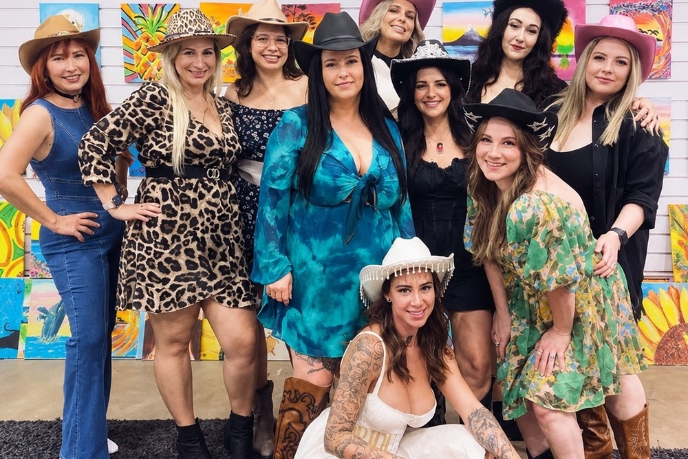 A cowgirl hens crew in hats posing in front of a wall of painted canvases