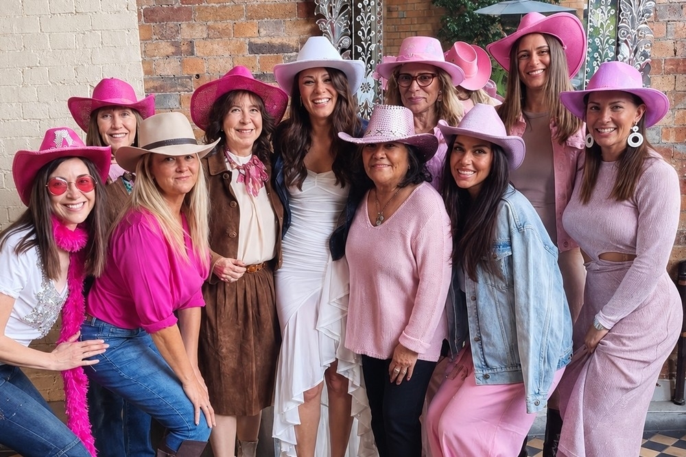 Hens of all ages in pink cowgirl hats smiling together in a brick courtyard
