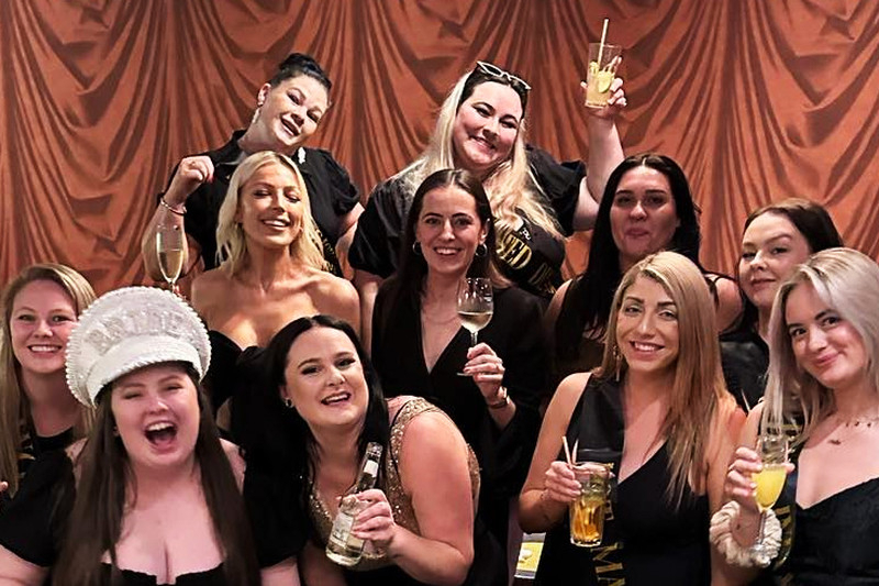 A hens group in black dresses raises drinks in a red velvet lounge