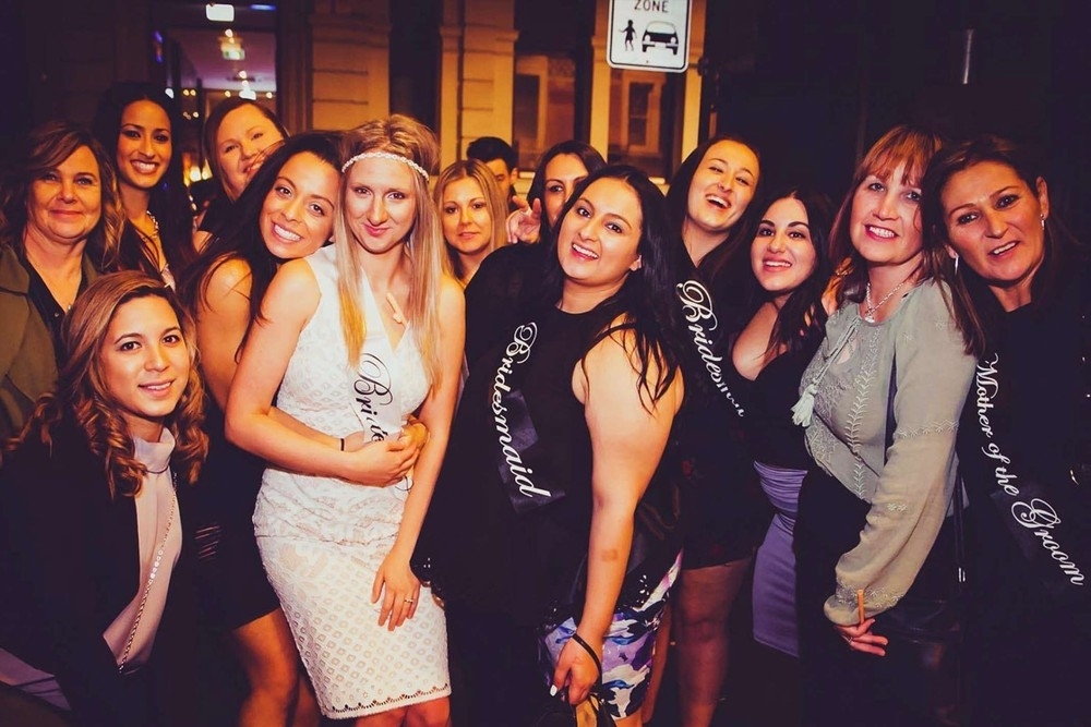 A bride in white and her friends in sashes smile together on a night out