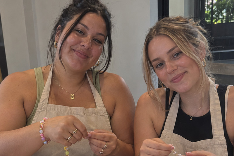 Two friends in aprons hold up the charm bracelets they are threading
