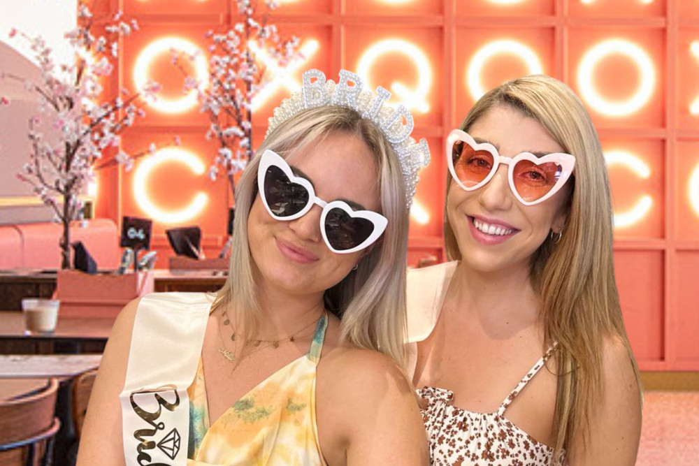 The bride in a sash and a friend in heart sunglasses in a pink neon room