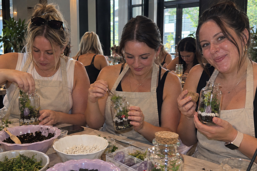 Three friends in aprons planting tiny gardens in glass jars at a workshop table