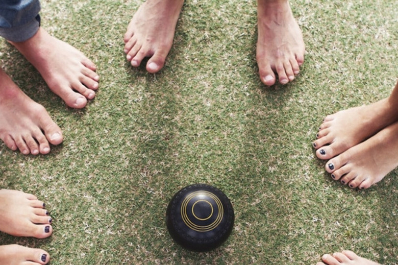 Bare feet with painted toenails standing around a black lawn bowl on the grass