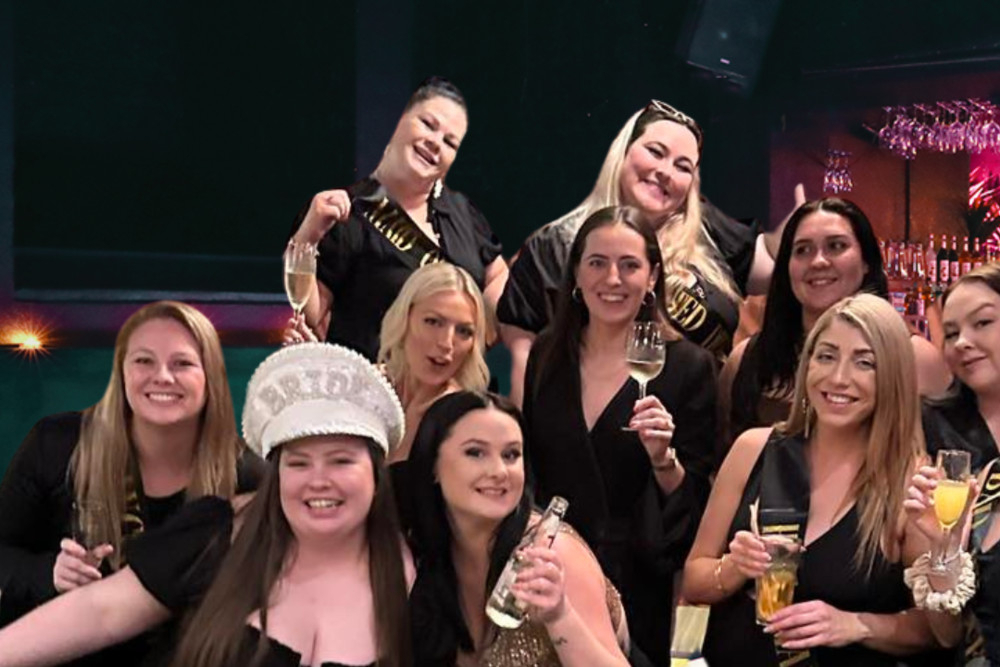 A big hens crew in black crowded together at a bar, the bride in a white sequin cap