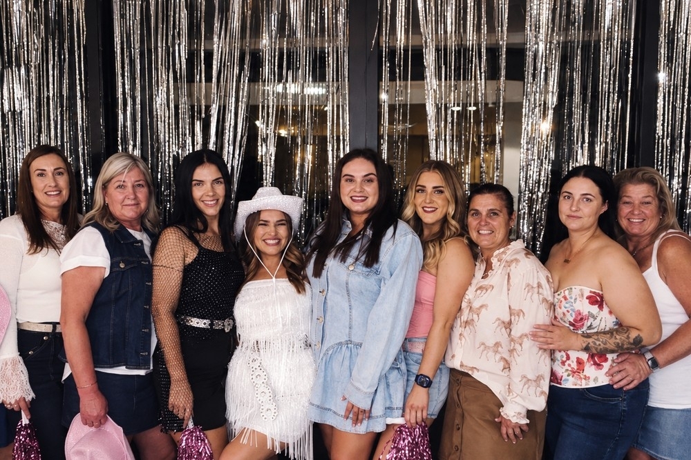 A cowgirl hens group of all ages with the bride in a white hat in front of silver tinsel