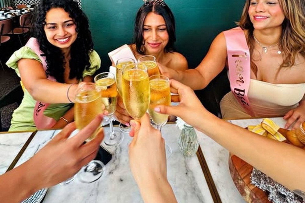 Hens in sashes and a tiara clinking glasses of bubbly across a marble table