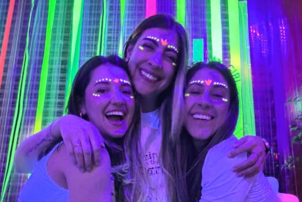 Three friends with glowing face paint hugging and laughing under UV light