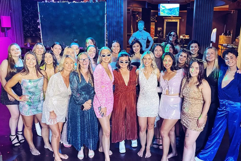 A big hens crew in sequins and party dresses lined up at our male revue show, with a performer on the lit stage behind them