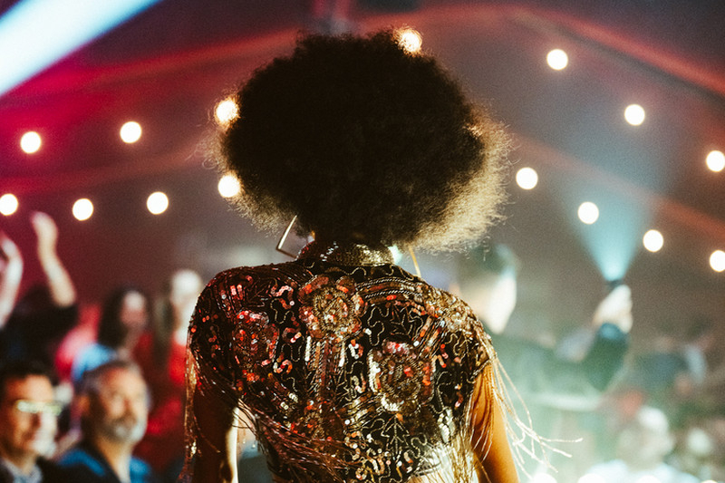 A performer in a sequinned fringe jacket walking past a seated audience under stage lights