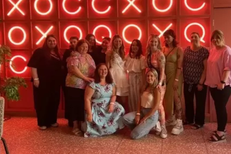 A hens crew posing in front of a glowing wall of red neon letters