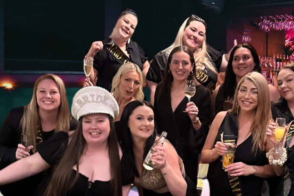 A hens group dressed in black around the bride in a sparkly hat, glasses raised