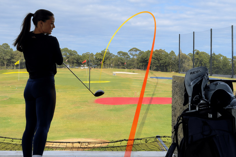 A woman driving a golf ball down the range, its flight traced in orange