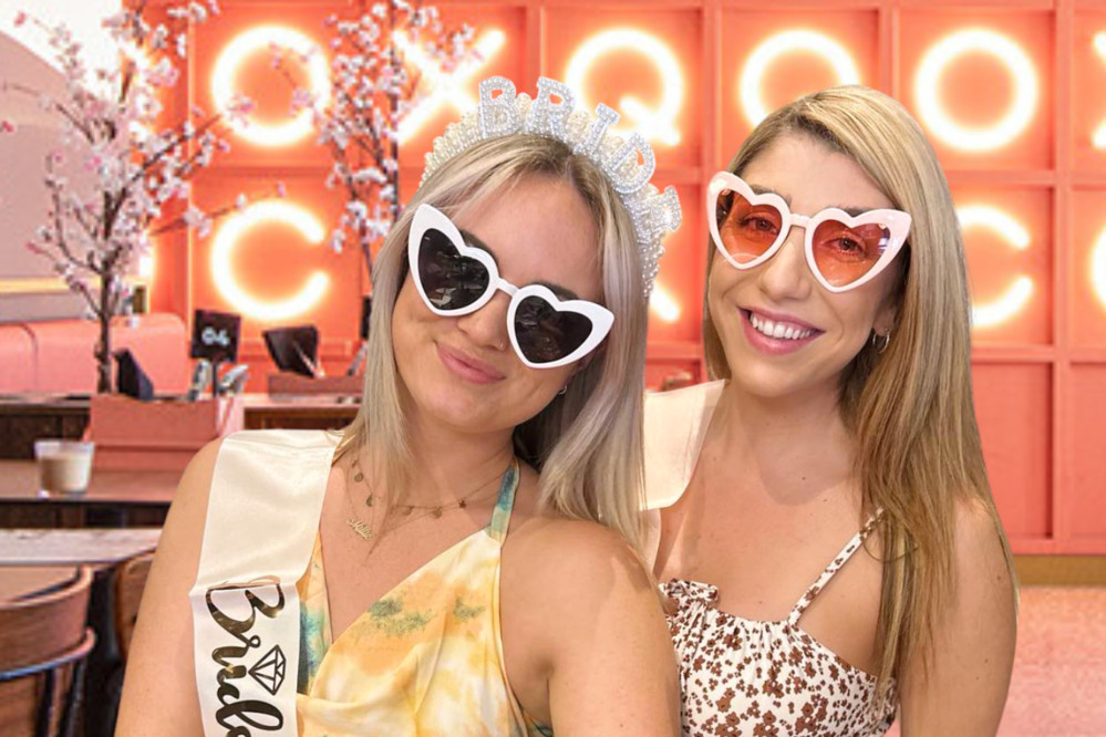 The bride in a sash and heart sunglasses cheek to cheek with a friend under pink neon letters
