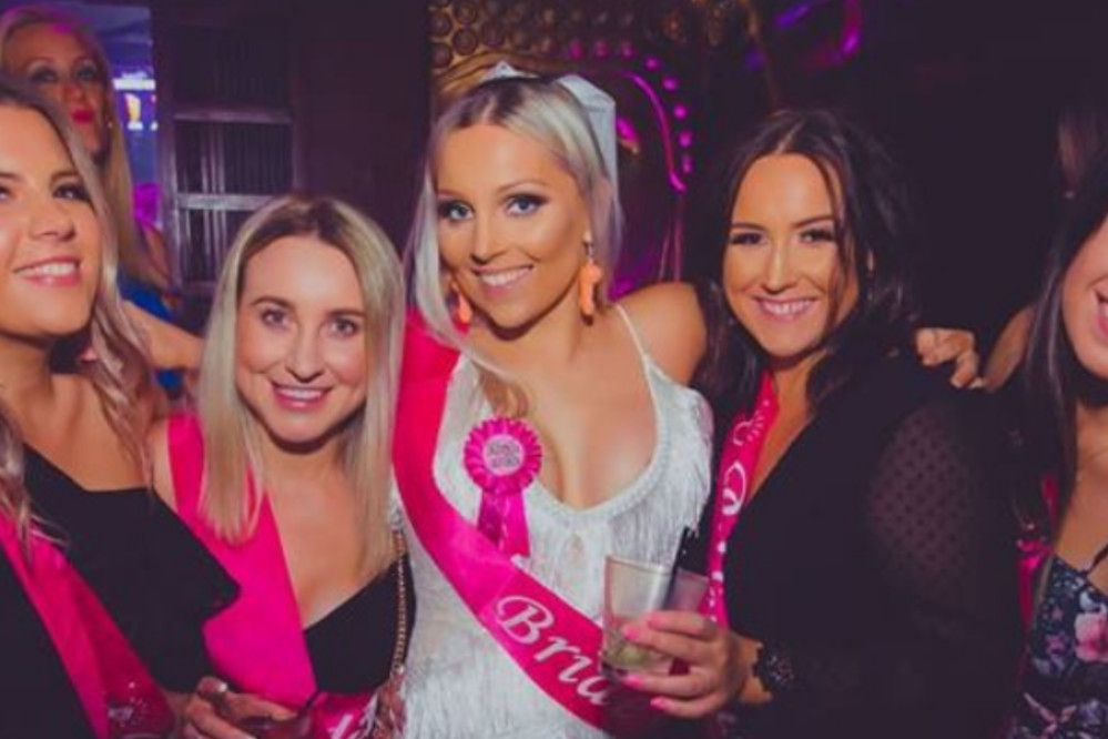 The bride in white with her friends in pink sashes, all smiling together in a lounge