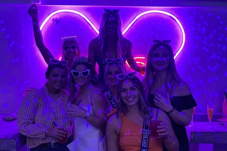 A hens crew in heart sunglasses posing under a glowing purple neon heart
