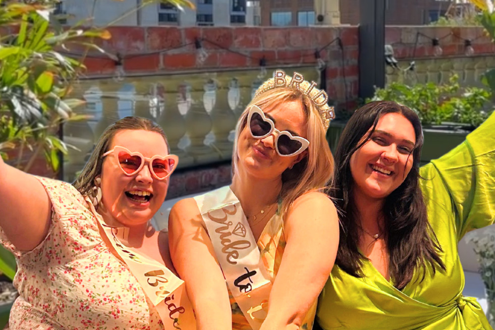 A bride to be in a sash and heart sunglasses with two friends on a sunny rooftop
