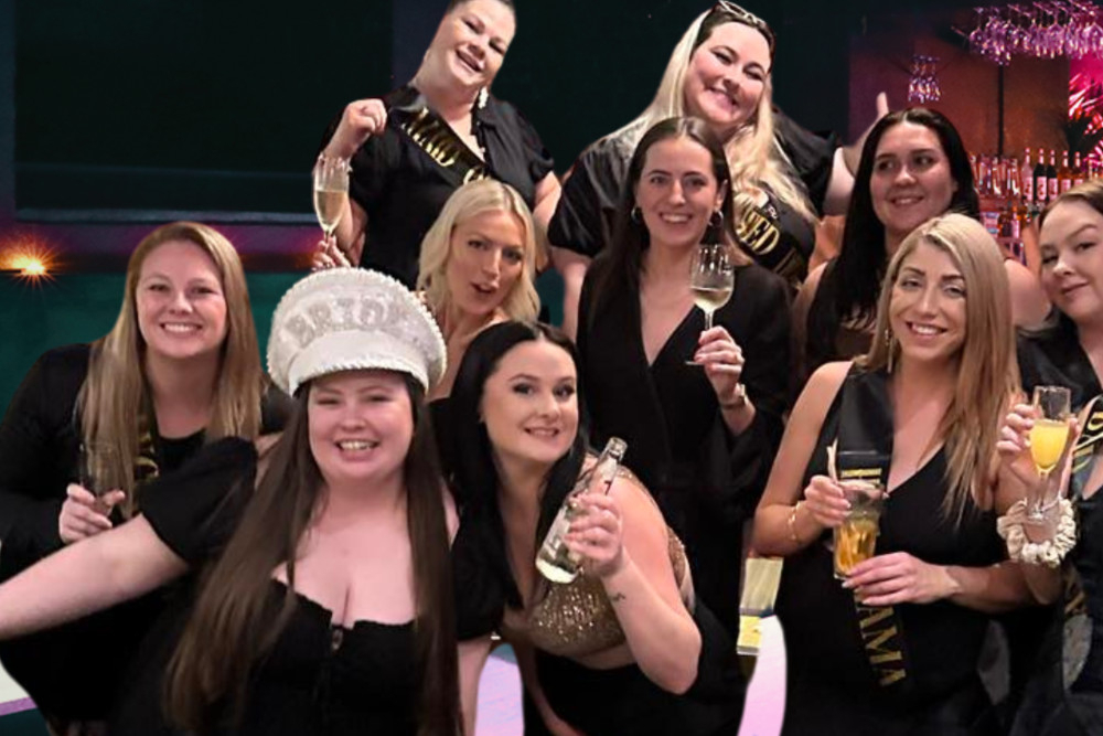 A hens group in black dresses around the bride in a sparkly cap, drinks raised