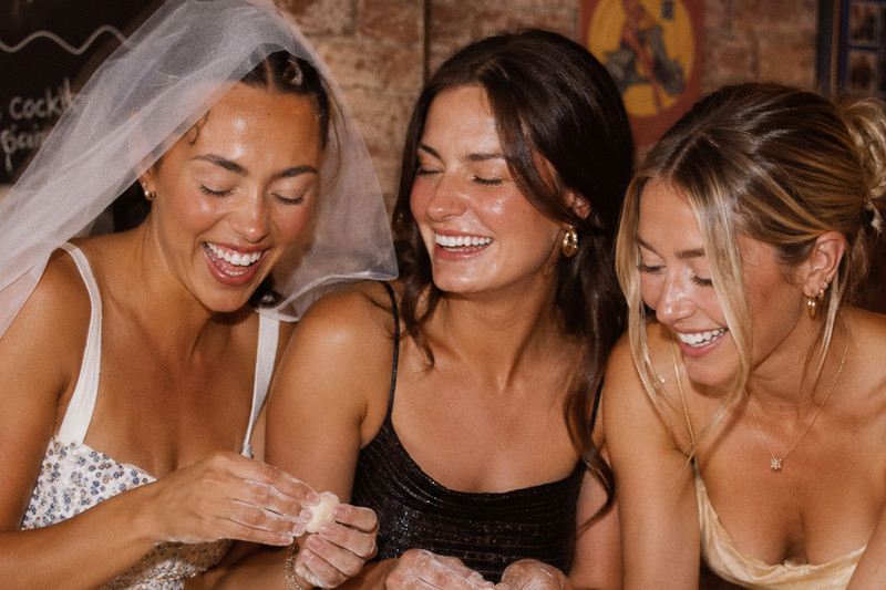 A bride in a veil laughing with two friends as they roll gnocchi by hand at a hens cooking class