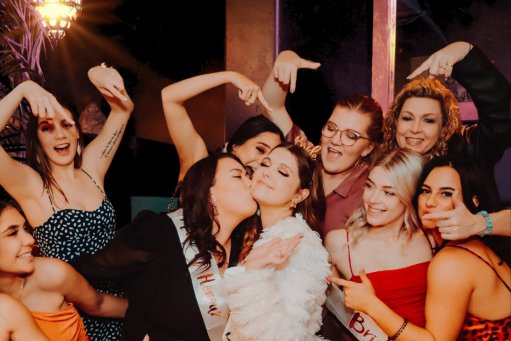 Hens dancing and laughing around the bride in white as one friend kisses her cheek