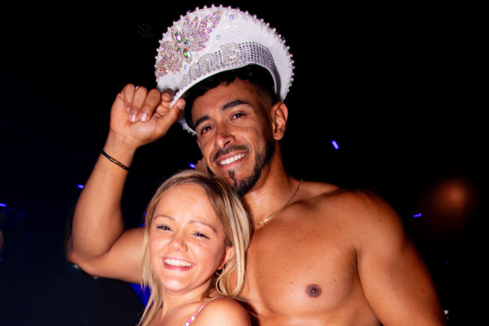 A laughing hen in pink with a shirtless waiter wearing a sparkly bride cap