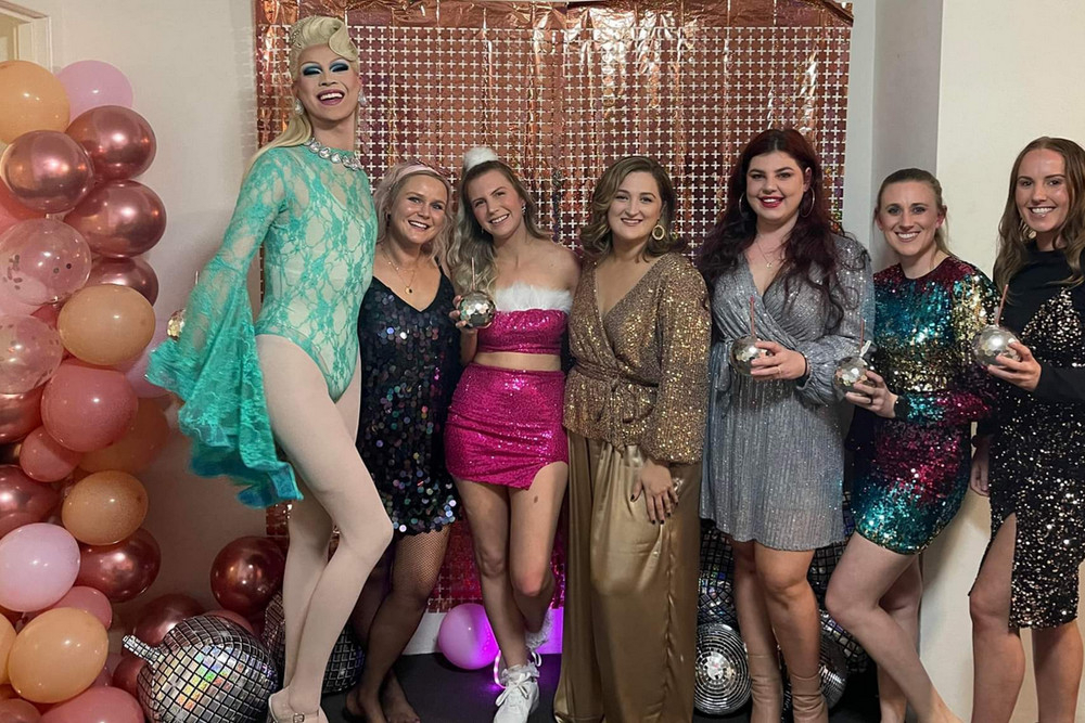 A drag queen in teal lace with a hens group in sequins beside a balloon garland